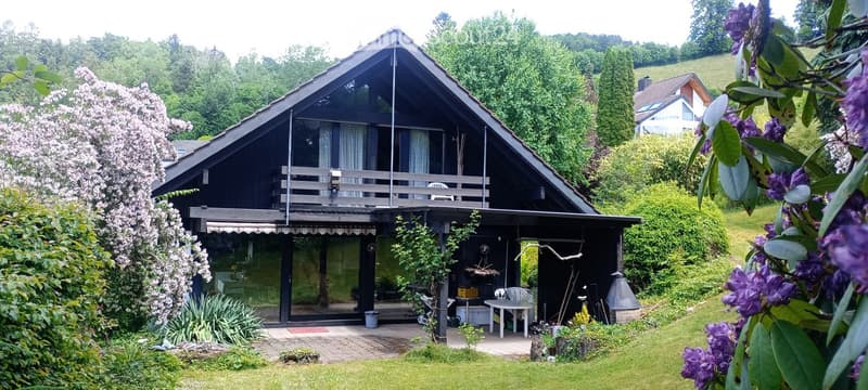 Two-story house with a dark wooden exterior, large windows, and a balcony on the upper level. The surrounding area includes a well-maintained garden with flowering trees and shrubs.