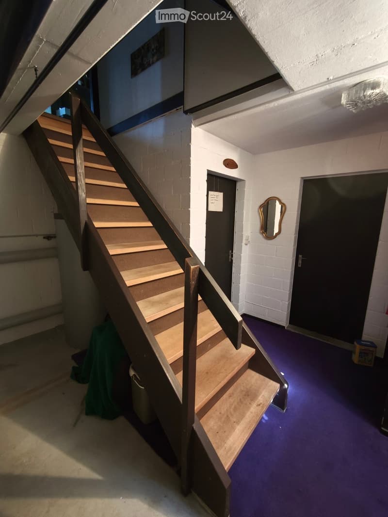 Staircase with wooden steps and metal railing, leading to an upper level. The hallway has white brick walls, a purple carpeted floor, and a small mirror on the wall. There is a green cloth draped over a surface near the stairs.