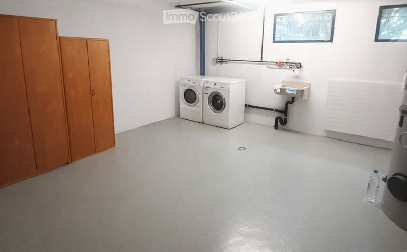 A clean, unfurnished utility room with a washing machine, a wooden cabinet, and a small sink. The room has white walls, a grey floor, and a window providing natural light.