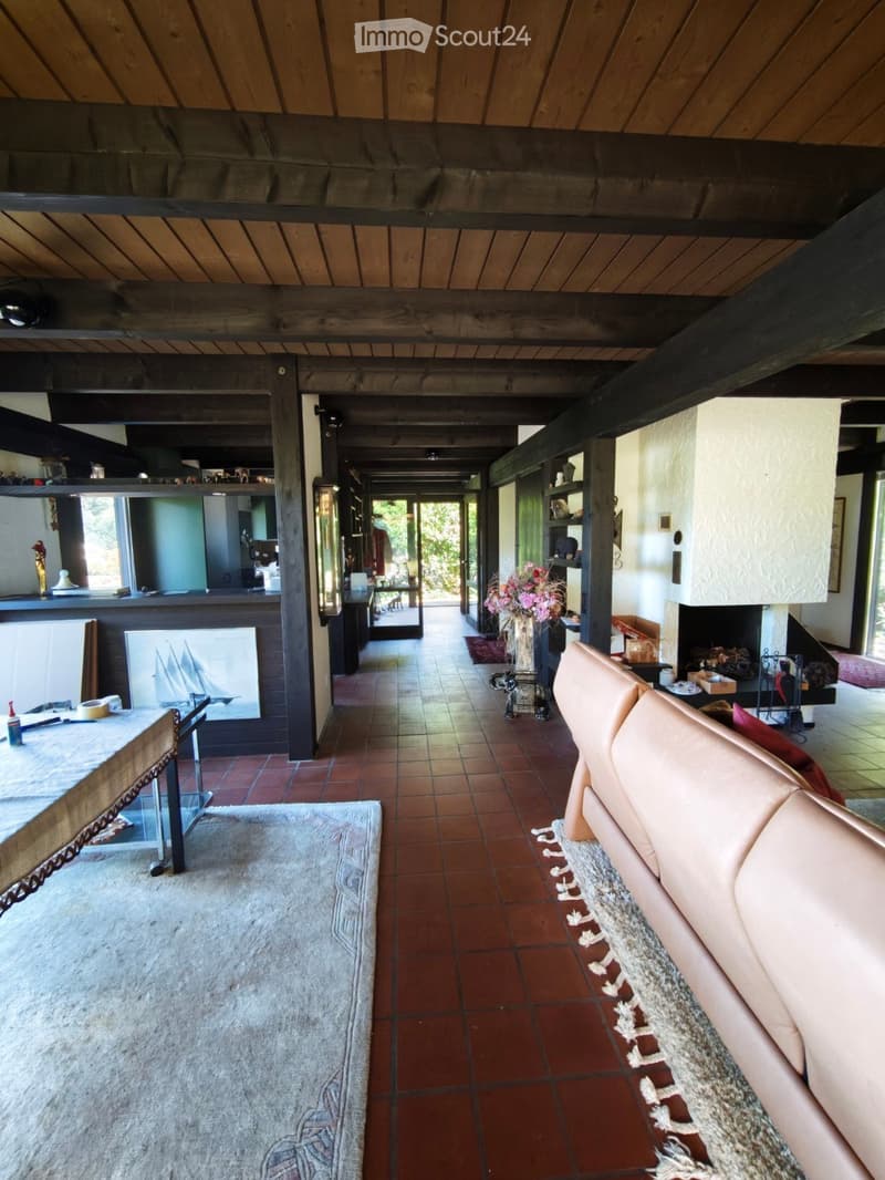 Open-plan living area with exposed wooden beams, terracotta tile flooring, a cream-colored sofa, a coffee table, a vase with flowers, and shelves with decorative items.