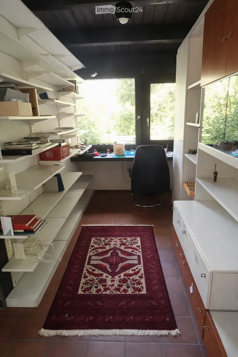 Bright room with wooden floor, red patterned rug, white shelves filled with books and items, a desk with a chair, and a large window with a view of greenery.