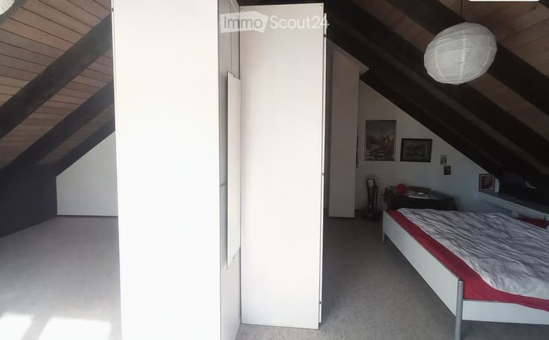 Modern attic bedroom with slanted wooden ceilings, white walls, a bed with red and white bedding, a nightstand, and several picture frames on the wall.