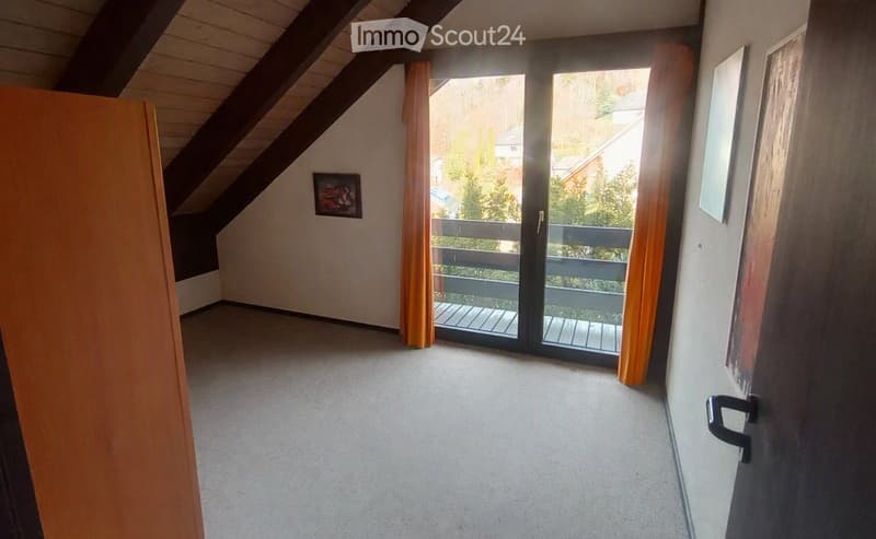 An empty attic room with a sloped wooden ceiling, carpeted floor, and a large sliding glass door leading to a balcony with a wooden railing and a view of greenery.