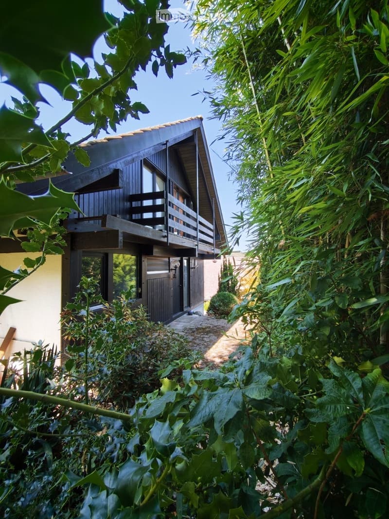 A two-story house with a wooden exterior, a balcony on the upper level, and a pathway leading to the entrance, surrounded by lush greenery and plants.