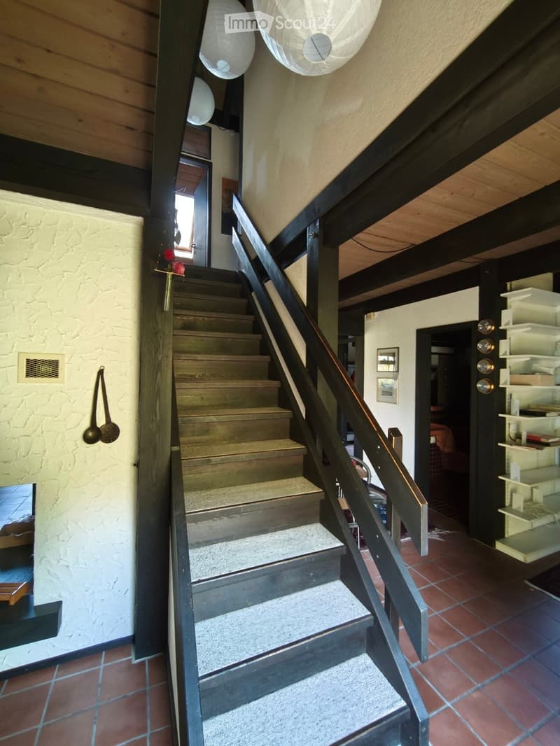 Staircase with wooden steps and black railing, leading to an open area with exposed wooden beams and white paper lanterns hanging from the ceiling. The walls are textured, and the floor is tiled.
