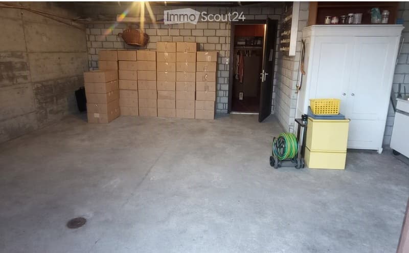 Empty concrete garage with exposed brick walls, stacks of cardboard boxes, a white cabinet, and a coiled green hose on a cart.