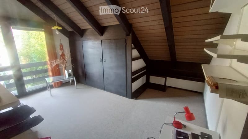 Modern, open-concept room with exposed wooden beams, a staircase leading upwards, a glass table with a chair, a tall cabinet, and a red lamp on a counter.