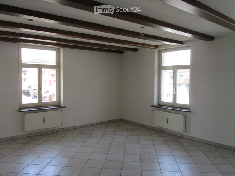 Empty room with white walls, tiled floor, wooden beams on the ceiling, and two windows with white frames.