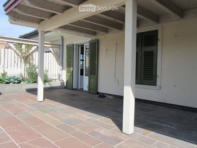A covered patio with brick flooring, white columns, and green shutters. The patio is adjacent to a white fence and a white wall.