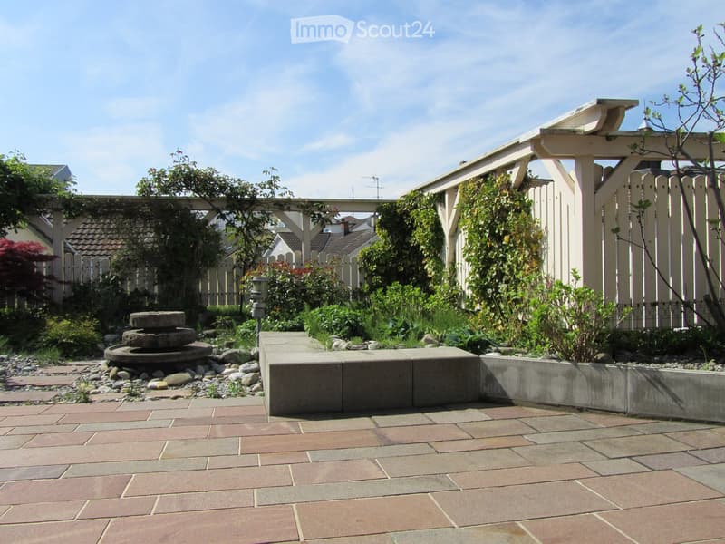 A garden area with a brick floor, a fountain, and a white wooden fence. The garden is surrounded by plants and trees.
