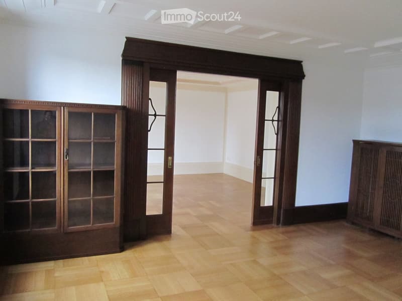 Empty room with wooden parquet flooring, white walls, and two wooden cabinets with glass doors. The room has a large open doorway leading to another room.