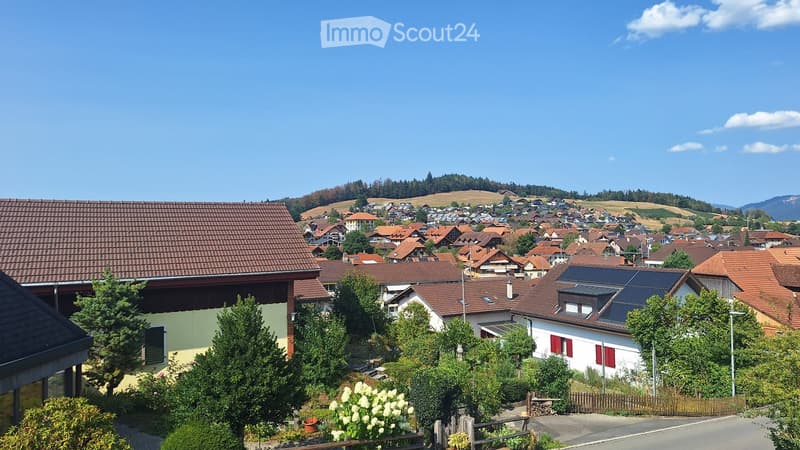 A picturesque village with red-tiled roofs, surrounded by lush greenery and hills under a clear blue sky.