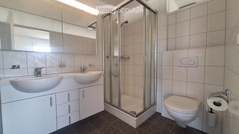 Modern bathroom with two sinks, a large mirror, a walk-in shower, and a toilet. The space features white tiles and a dark brown floor.