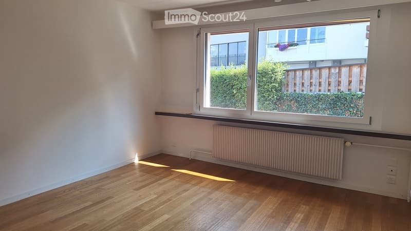 Empty room with wooden flooring, white walls, large window with view of greenery, radiator below window, and electrical outlets on walls.