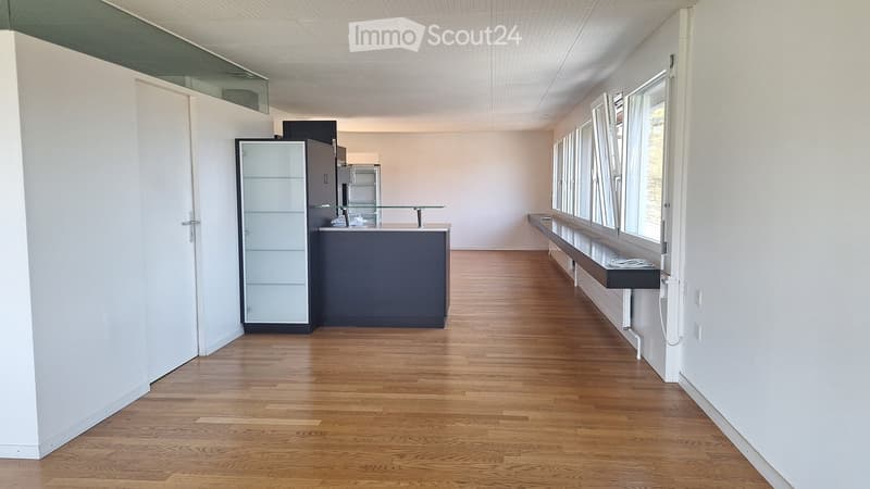 Empty room with light-colored walls, wooden flooring, and large windows. Features a glass-fronted cabinet and a small counter with a glass top.