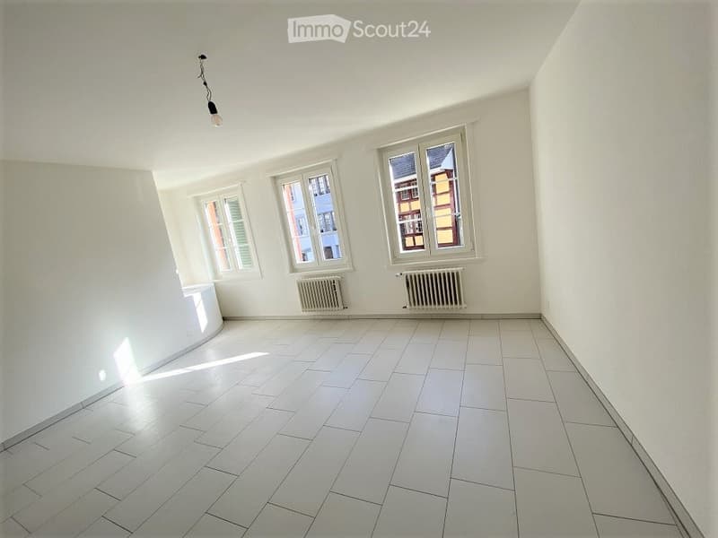 Empty room with white tiled flooring, white walls, three windows, radiators, and a single hanging light fixture.