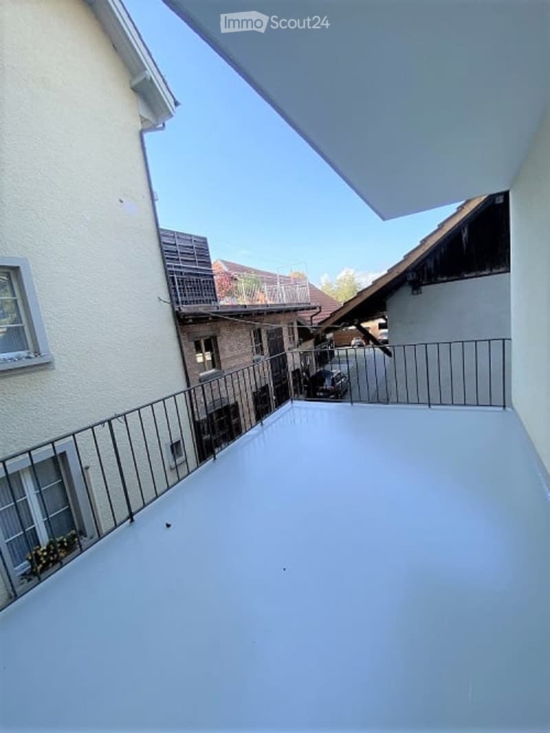 Balcony with metal railing, white floor, overlooking a street with neighboring buildings and parked car.
