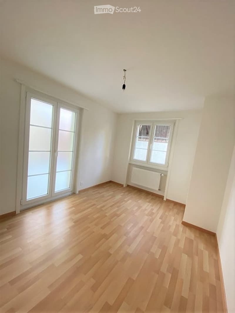 Empty room with light wooden flooring, white walls, large windows, and a radiator. No furniture or equipment present.
