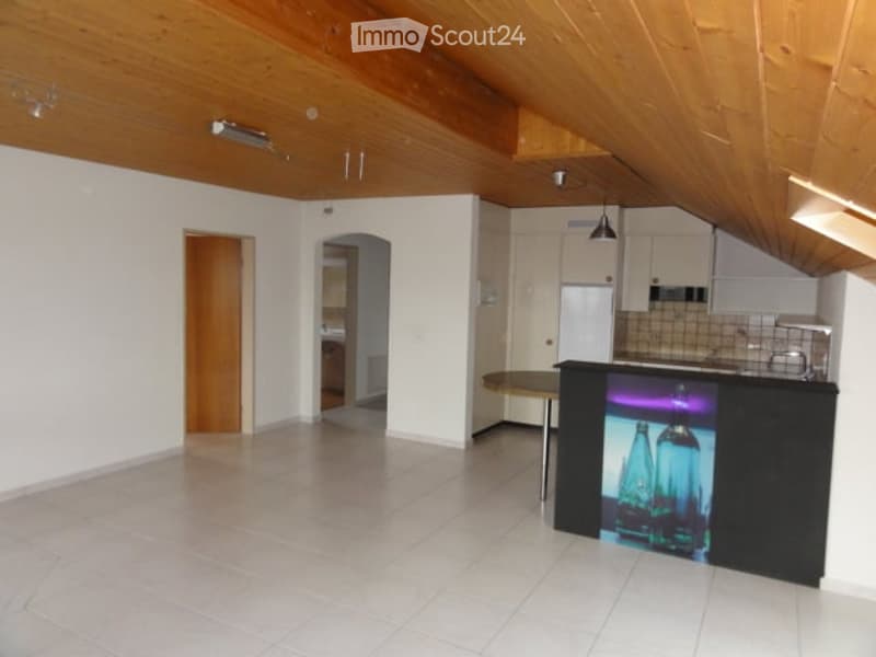 Open-plan kitchen and living area with white tiled flooring, wooden ceiling beams, and white cabinetry. The space is unfurnished, showing only structural elements like walls, doors, and built-in cabinets.