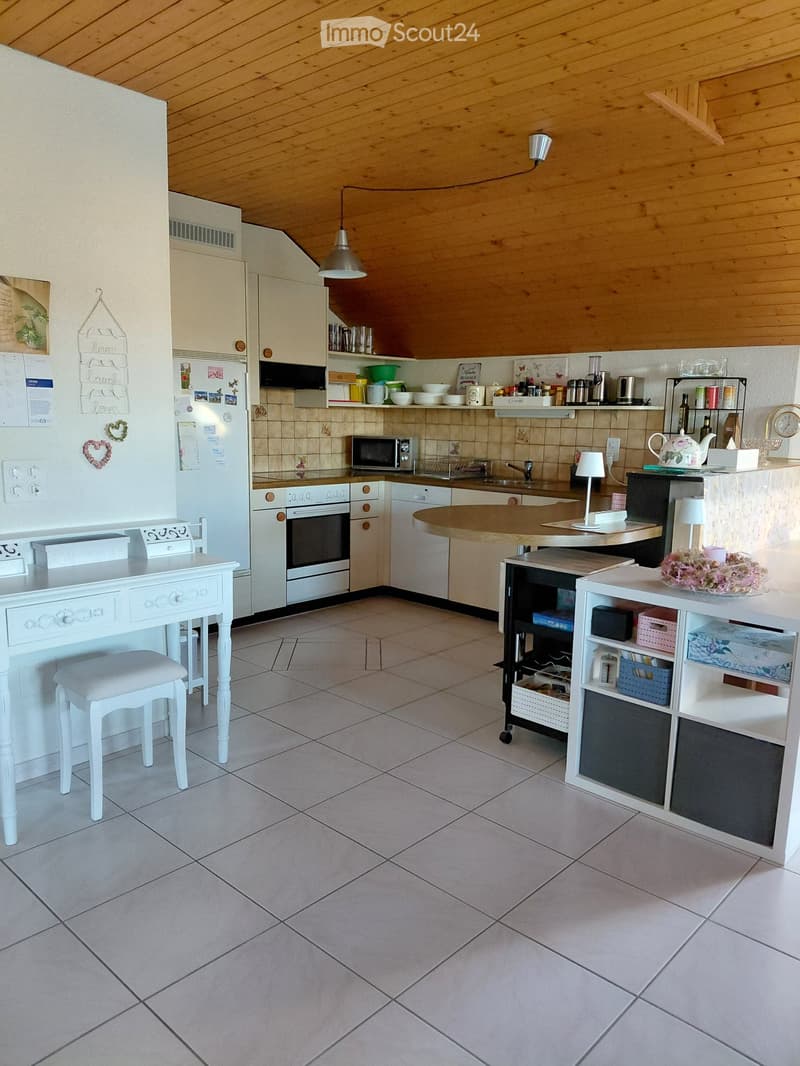 Bright kitchen with white cabinets, tiled walls, and a central island. Includes a refrigerator, oven, microwave, dishwasher, a small table with a stool, and various kitchen items on the counters.