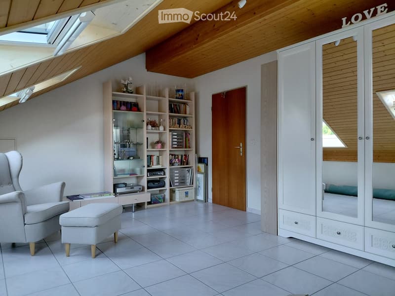 Bright, modern room with a slanted wooden ceiling, large skylight windows, a white armchair, a matching ottoman, a bookshelf filled with books and decorative items, a mirror cabinet, and a small table with books and a printer.