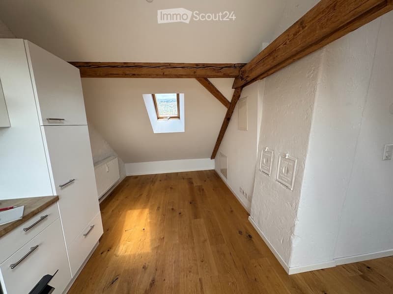 An unfurnished attic room with wooden floors, exposed wooden beams, and a skylight window. The room features white cabinetry on one side and a small staircase leading to another area.