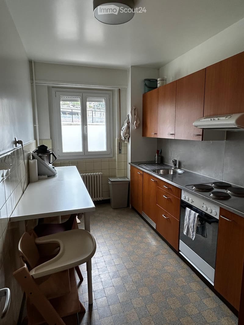 A small kitchen with wooden cabinets, a white countertop, a sink, a stove, and a small table with a chair. The room has tiled walls and a tiled floor, with a window providing natural light.