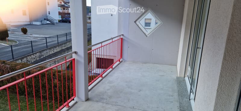 Balcony with red metal railing, concrete floor, and a view of a parking area and building.