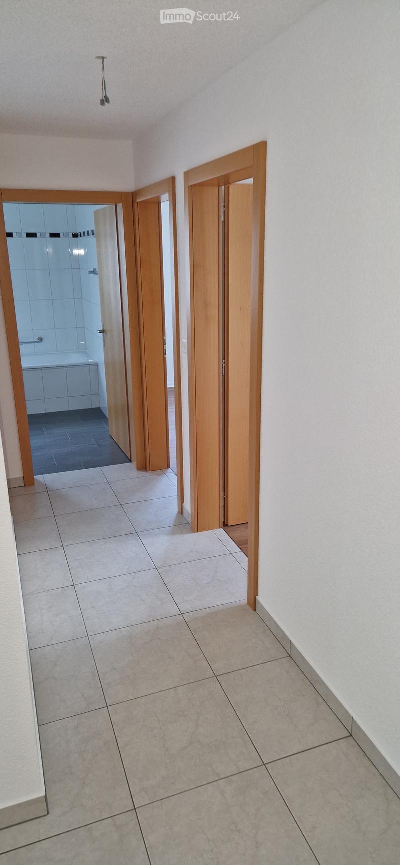 Empty hallway with tiled floor and white walls, featuring two wooden doors and an open doorway leading to a tiled bathroom.