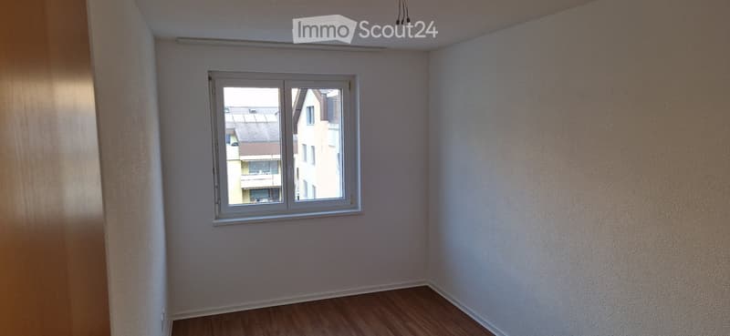 Empty room with wooden flooring, white walls, and a single window showing a view of neighboring buildings.
