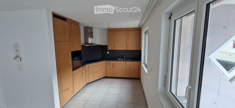 Modern kitchen with light wooden cabinets, black countertops, and stainless steel appliances, including a sink, faucet, oven, and range hood. The space features tiled flooring and a window with a view.