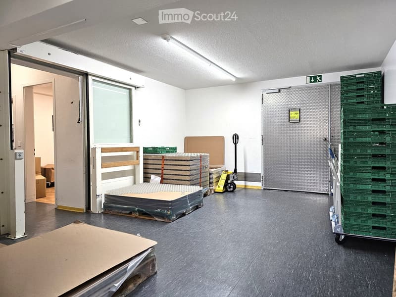 Empty storage room with grey flooring, white walls, and large industrial doors. Stacks of cardboard boxes and green crates are visible, along with a yellow pallet jack.