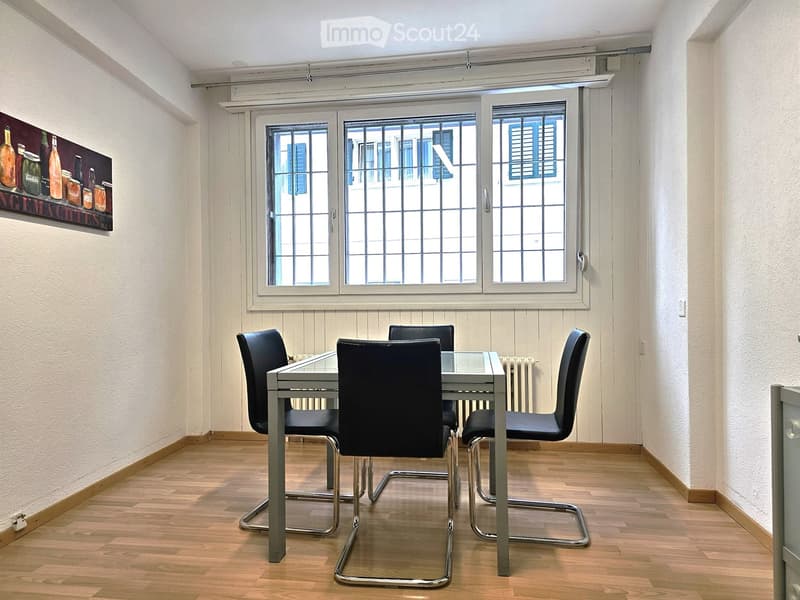 Dining room with a glass table and four black chairs, wooden flooring, and large windows with shutters.