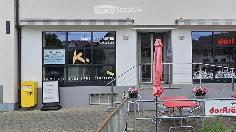 Facade of a commercial building with large windows, signage for "Konfi Manufaktur Markus Kunz" and "LA VIE EST DURE SANS CONFITURE", outdoor seating area with tables and chairs, and a yellow post box.