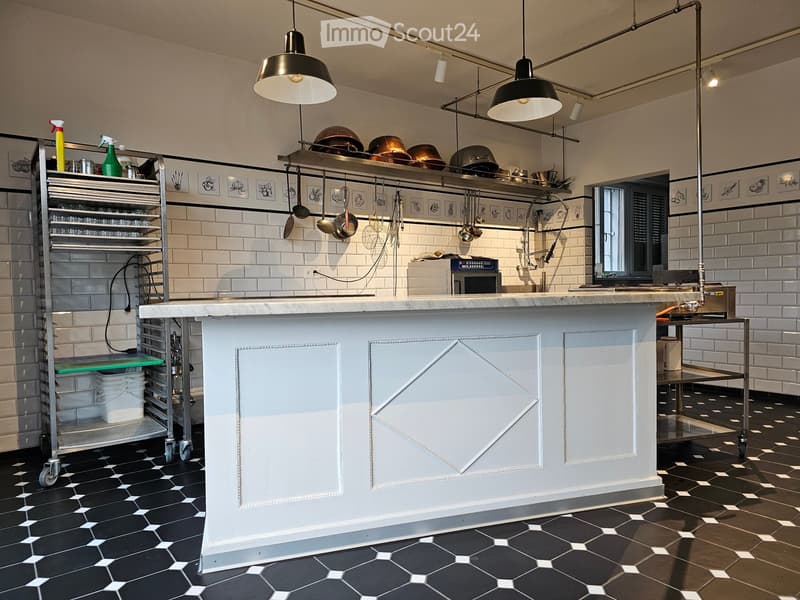 Commercial kitchen with white brick walls, black and white hexagonal tile floor, stainless steel counter, hanging pots, and pantry cart.
