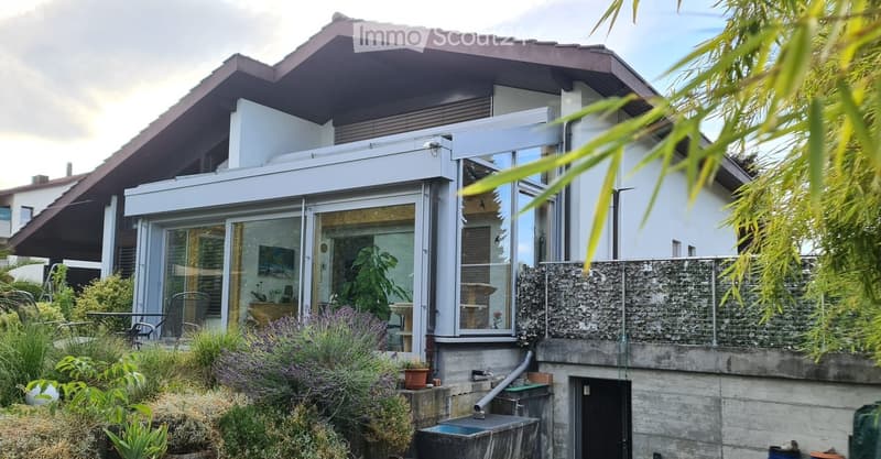 Modern two-story house with large glass windows and a balcony, surrounded by lush greenery and plants.