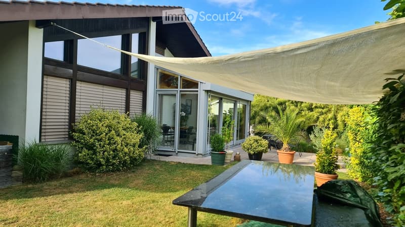 Garden area with a modern house in the background, featuring a patio with a glass table, potted plants, and a large sunshade. The house has large windows and a sliding glass door leading to the patio.