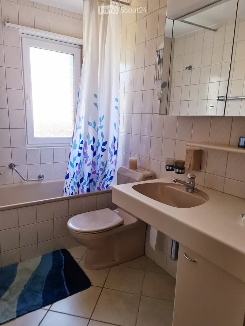 Bathroom with a sink, toilet, bathtub, and a mirror. Features a shower curtain with a leaf pattern, a towel on the floor, and a candle on the sink counter.