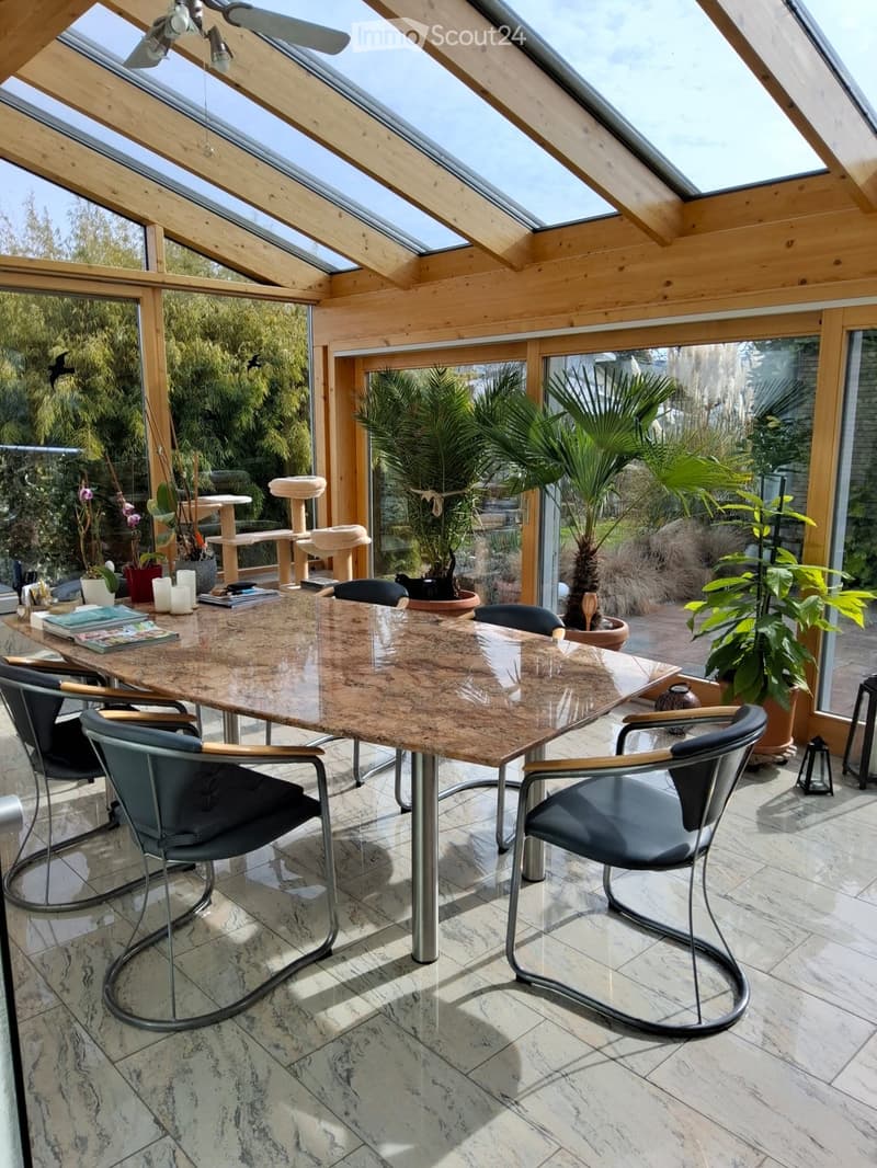 Brightly lit conservatory with a large marble dining table, four modern chairs, a cat tree, potted plants, and decorative candles.