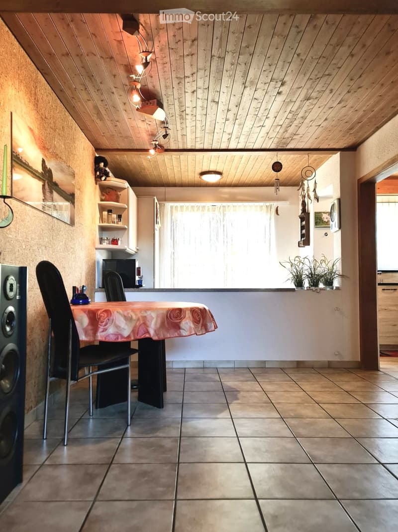 Dining area with a table, chairs, shelves, and a refrigerator in the background. Wooden ceiling and tiled floor.