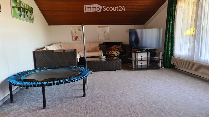 Living room with a sofa, coffee table, television, wicker chair, and a small treadmill. The room has a slanted wooden ceiling and large windows with curtains.
