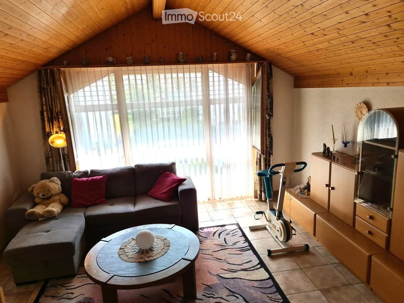 Cozy living room with wooden ceiling, large window with sheer curtains, grey sectional sofa with red pillows, coffee table with a decorative doily, exercise bike, and a mirror cabinet.