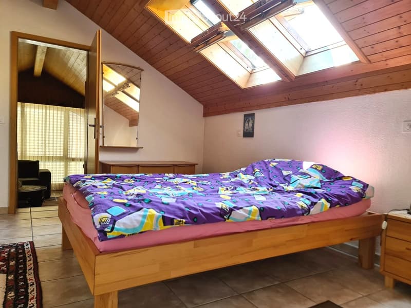 Cozy attic bedroom with wooden slanted ceilings, skylights, a double bed with colorful bedding, a nightstand, a small chair, and a mirror. The room features tiled flooring and a small rug.