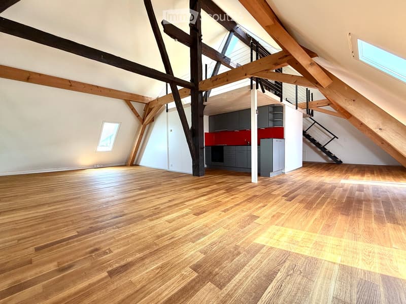 Open-plan attic space with exposed wooden beams, hardwood flooring, and a small kitchen area with dark cabinets and a red backsplash.