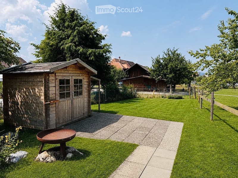 Well-maintained garden with a wooden shed, a metal fire pit, a concrete pathway, and a fenced area with trees and a house in the background.