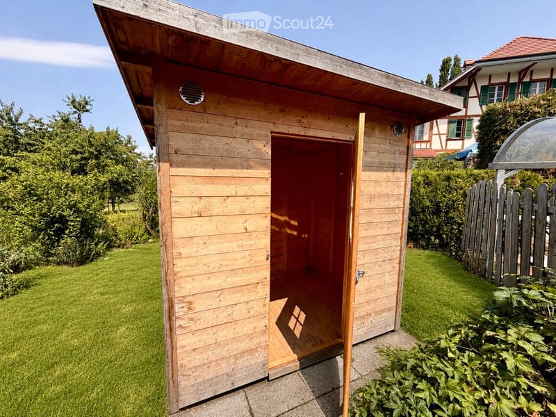 A wooden shed with an open door, situated in a grassy yard with surrounding greenery and a wooden fence.