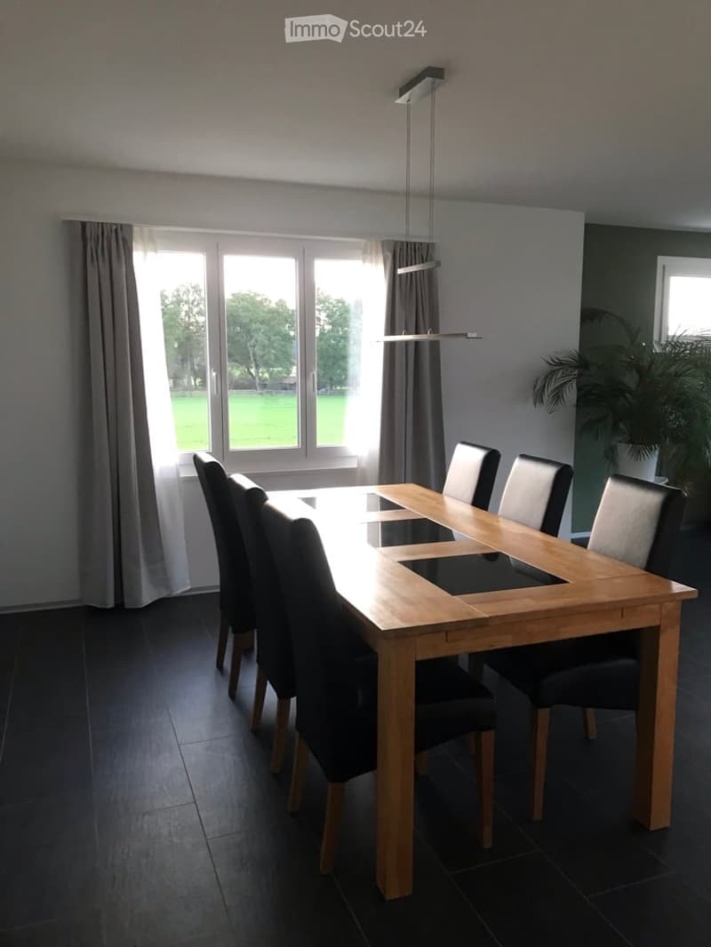 Modern dining room with a wooden dining table and black chairs, large windows with curtains, and a pendant light fixture.