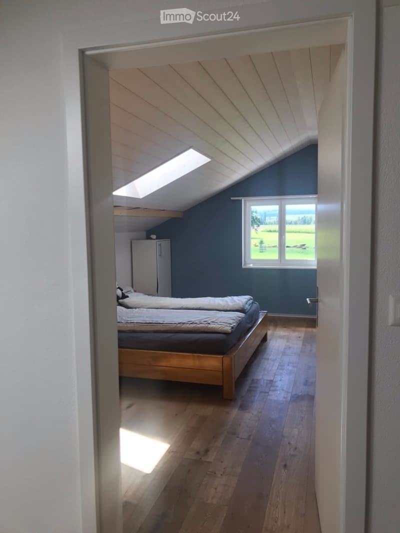 Bright attic bedroom with wooden flooring, slanted ceiling, skylight, and large window overlooking a green field.