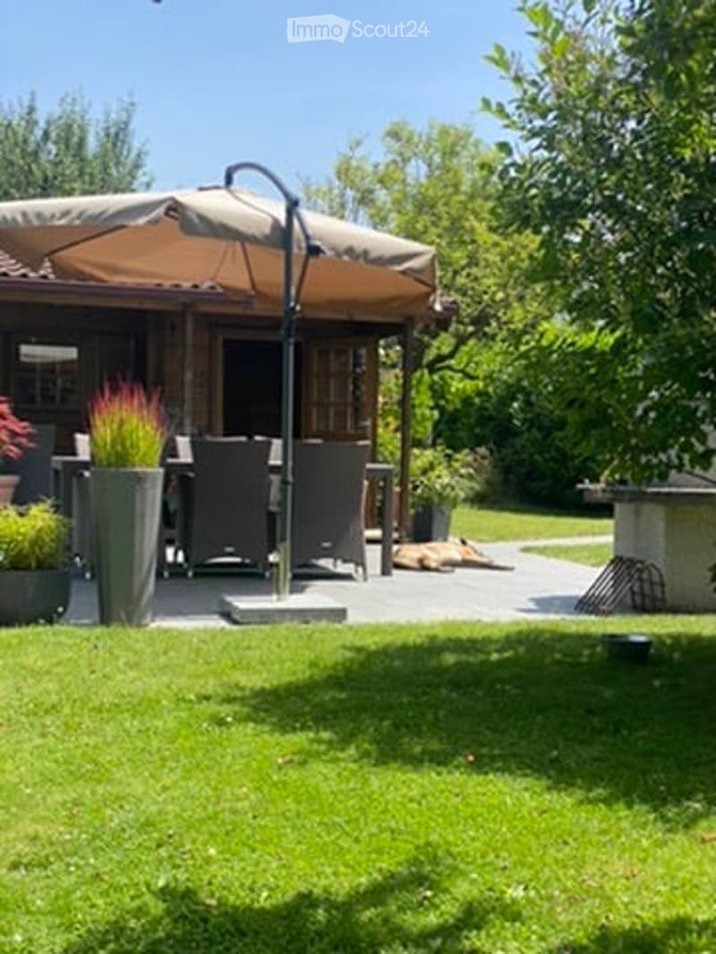 A well-maintained garden with a patio area featuring a large umbrella, outdoor furniture, and potted plants. The space is surrounded by greenery and includes a small dog lying on the grass.