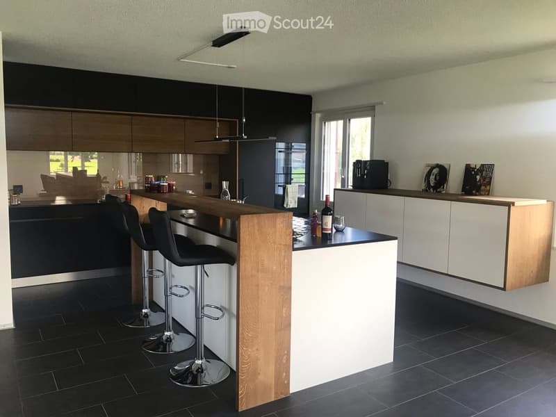 Modern kitchen with dark cabinets, wooden island, black stools, and pendant lights.